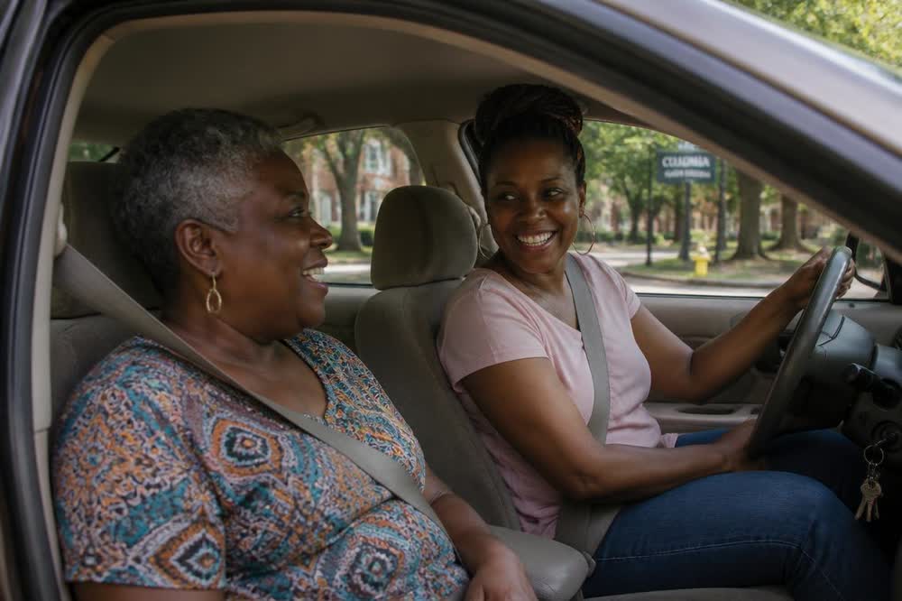 A Senior Express driver and her passenger talking and laughing together in the front seats of the car on a leafy Columbia street.